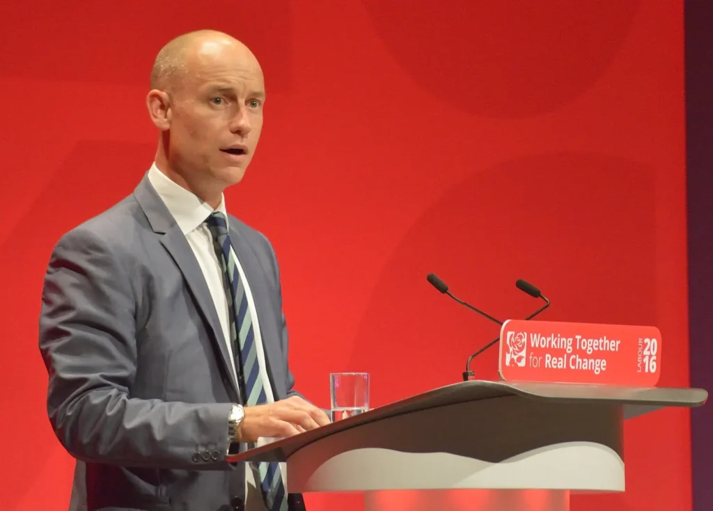 UK AI Clinical Trial Reforms Cut Approval Times in Half 1 UK Health Minister Stephen Kinnock and MHRA Chief Lawrence Tallon stand at a government press conference podium. A screen behind them displays a graph showing clinical trial approval times dropping from 91 to 41 days.